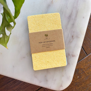 A pack of three yellow sponges, wrapped in brown paper rest on a granite slab.